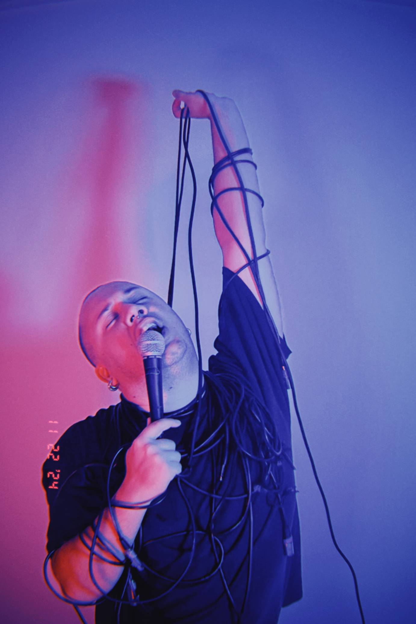 manin black t shirt wrapped in microphone cord, speaking into mic but looking also as if the mic cords are a noose with which he is hanging himself 